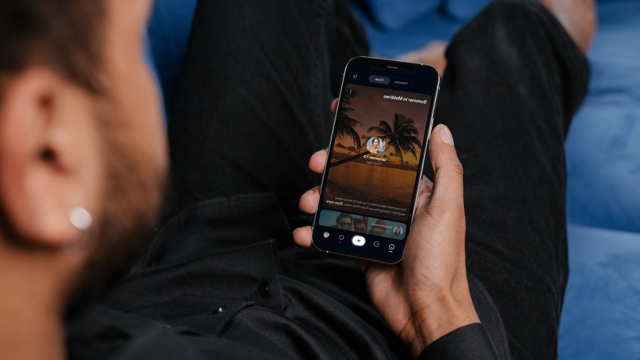 A person holding a smartphone showing a sunset memory on the Recap app while relaxing on a blue couch.